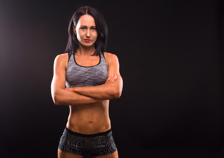 Image of beautiful fitness lady posing with her arms crossed or folded isolated on black background. Pretty brunette lady looking at camera and smiling in studio.の写真素材