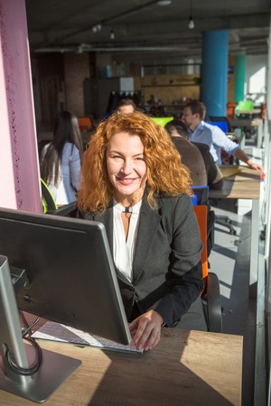 Portrait of happy smiling red haired businesswoman working on computer in office interior. Confident...
