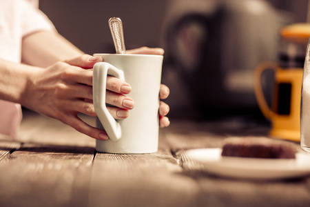 Female hands holding a white cup of tea standing close to wooden desk.の写真素材
