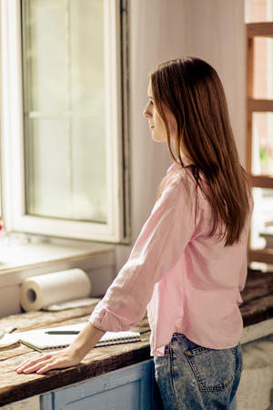 Rear view of young girl standing close to window and wooden desk.の写真素材