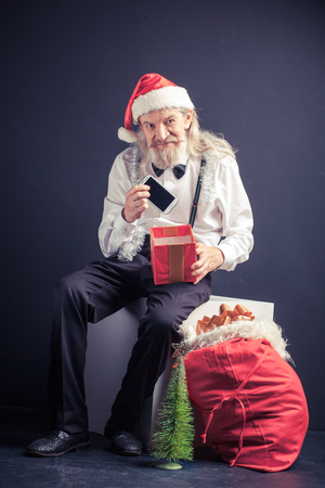 Happy office Santa holding cell phone as christmas gift. Old man in Santa hat pulling cell phone from gift box.の写真素材