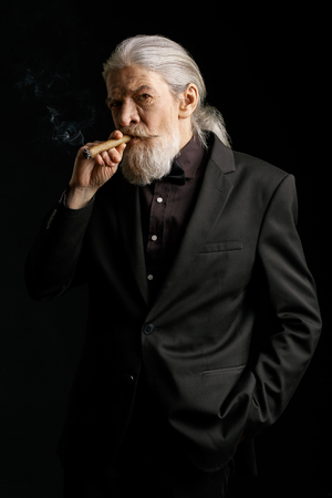Aged wrinkled man standing against black background. Grey haired male smoking cigar.の写真素材