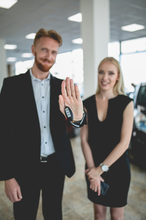 Beautiful couple in vehicle showroom holding keys from new car. Man and woman choosing new electro car. Image with selective focus.の写真素材
