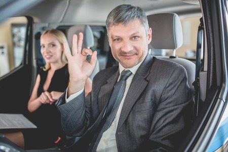 Vehicle dealer showing new car to pretty woman. Smiling manager sitting on front seats in new car showing ok with fingers.の写真素材