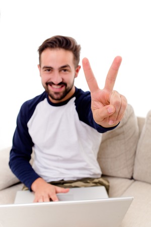 Male smiling showing V with fingers. happy young guy sitting on sofa. Image with selective focus.の写真素材