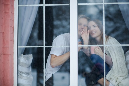 Attractive couple stay at the window. A girl is trying to show a boy falling snowflakes.