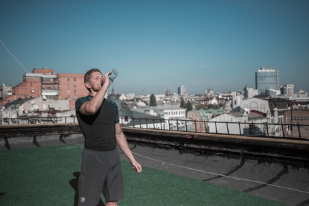 Sportive man drinking water after workout. Sprt on the roof of the building in the city, athletic male relaxing.の写真素材