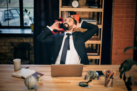 Mature businessman relaxing in armchair in luxury office. Talking to partner over smartphone.の写真素材