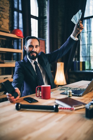 Nice man sitting at office and raising hand with money. Posing on camera.の写真素材