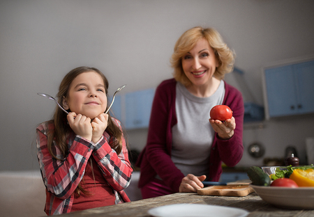 Grandmother and granddaughter posing on camera. Granny showing tomato and girl palying with spoons.の写真素材