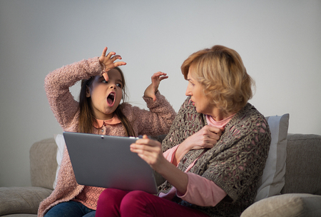 Little girl and granny playing on sofa. Girl scaring granny by holding hands. Granny holding tablet.の写真素材