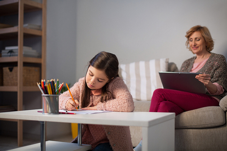 Pretty little girl sitting at small table and drawing. Her granny sitting on soaf with tablet.の写真素材