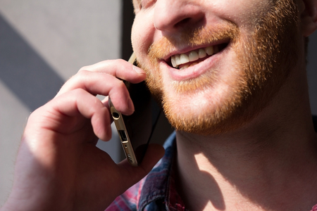 Guy having conversation on phone close up. Close up picture of man with ginger beard smiling holding mobile phone to his ear.の写真素材