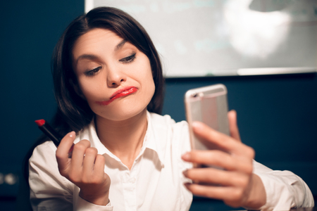 Lady taking selfie with messed up lipstick. Funny looking business woman smudging her lip make up purposefully while holding her phone in other hand.の写真素材