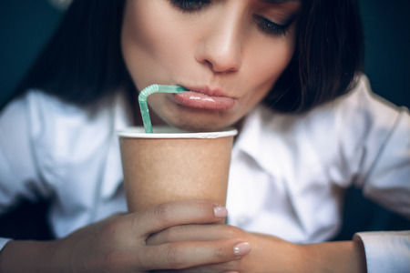 Pretty lady looking down in her coffee cup. Close up portrait of beautiful woman drinking coffee from green straw with her lips rolled. sの写真素材
