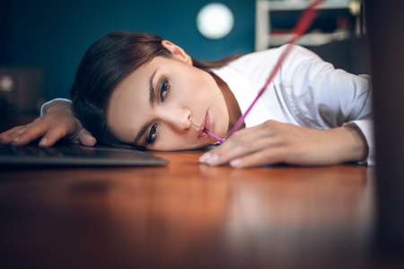Gorgeous lady drinking with long straw. Tired beautiful woman drinking from straw while lying on wooden office table with sad expression on her face.sの写真素材