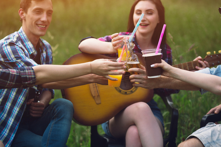 Three people toasting their beverages in park. Girl with guitar sitting in grass field next to two of her friends cheering drinks.の写真素材