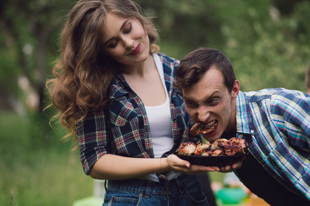 Young man trying to eat piece of chicken from plate of meat. Guy acting silly trying to steal piece of meat from bowl that girl is holding with both of her hands.の写真素材