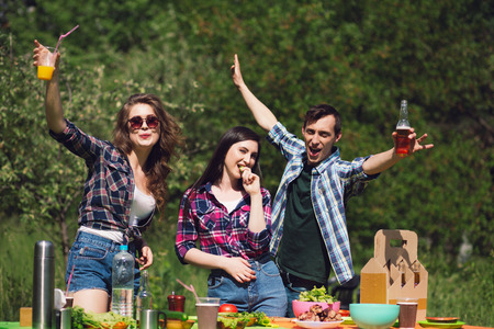 Three happy young people at picnic table. Excited friends standning behind picnic tablewith lots of food on it with drinks in their hands.の写真素材