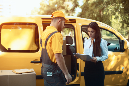 Lady signing off her delivery package. Delivery man wearing yellow shirt and hat and overalls waiting for client to put their signature.の写真素材