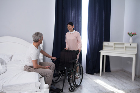Nurse bringing wheelchair to patient. Beautiful asian caregiver pushing wheelchair to bed on which elderly man is sitting.の写真素材
