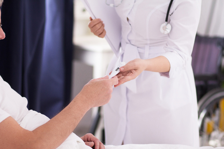 Female nurse giving her patient thermometer. Picture of doctor wearing white coat handing thermometer to patient in bed to measure his temperature.の写真素材