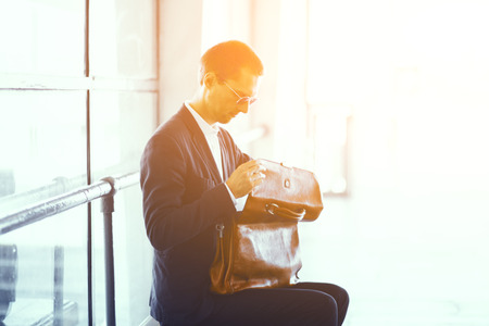 Handsome businessman sitting on bench with bag on his lap. Good looking man wearing elegant black suit and white collar shirt taking look inside his leather bag.の写真素材