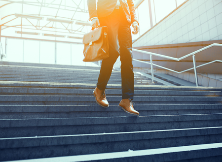 Legs of businessman walking down stairs. Cropped shot of man wearing suit and holding leather bag making his way down concrete stairs outdoors.の写真素材