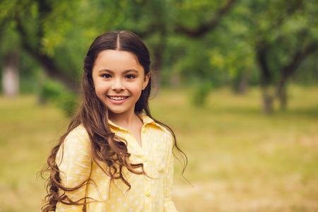 Portrait of smiling sweet little lady. Gorgeous little girl with long hair wearing yellow dress with puff sleeves smiling. Shot outdoors in forest or park.の写真素材