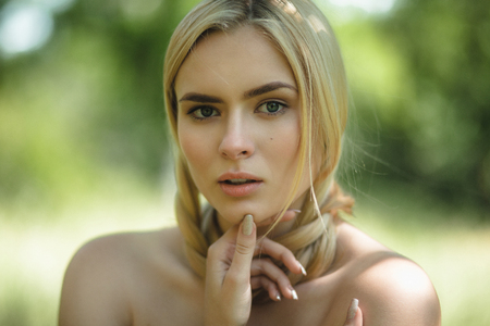 Blond lady with her hair wrapping her neck. Portarit of woman with bare shoulders posing outside in nature with her hand at her chin.の写真素材