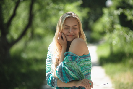 Charming young lady in park. Sweet blonde girl wearing knitted blue and purple sweater hugging herself and smiling with delight on her face.の写真素材