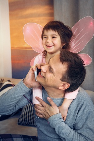 Father sitting on sofa in home clothes and speaking over phone. Little daughter with play wings hugging him.の写真素材