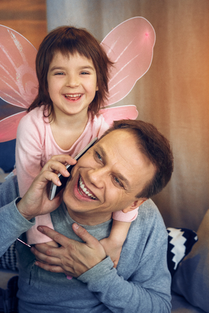 Daddy speaking over phone to colleague while little daughter having play wings hugging him.の写真素材