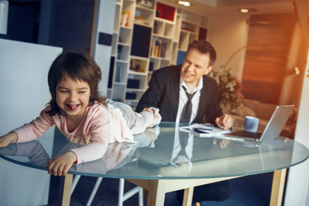 Little daughter lying on table and smiling while her father working on laptop. Business at home concept.の写真素材