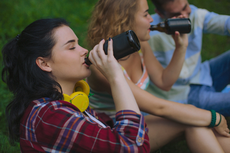 Group of students drinking in a park. Beautiful brunette girl drinks a beer in the park. Besties drink and enjoy their time together.の写真素材