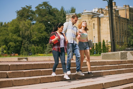 Happy students are walking together on campus. One man with two girls. Students between lectures getting sown the stairs in a parkの写真素材