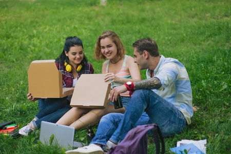 Group of students sit on the grass ans open the boxes with pizza. Pretty girl smiles and hold a box from pizza. Handsome man with a tatto looks into the box.の写真素材
