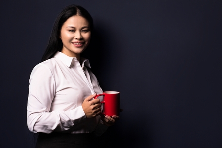 Businesswoman enjoying coffee break holding red cup. Standing half turn and smiling.の写真素材