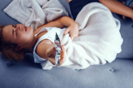 Close look on a womans hand holding a packs of pills. Blurred image of young blonde girl lying on the sofa feeling pain and holding blisters pf pills.の写真素材