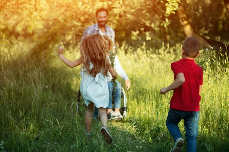 Little Girl Holds A Bouquet Of Wild Flowers And Runs To Her Woman Who Is Sitting In The Wheelchair Folloed By Cute Brother.の写真素材