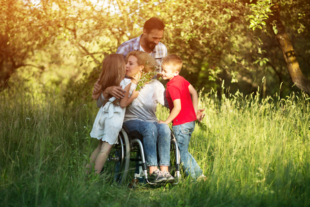 Cheerful Woman In Wheelchair Kisses Her Pretty Little Daughter And Hugs Son While Caring Man Looks Happily Over Themの写真素材