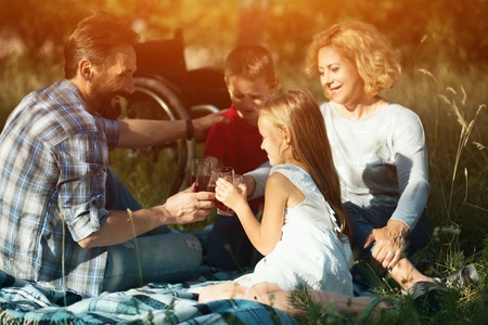 Attractive Father Of Two Kids Laughs With Children And Disabled Mother Is Sitting Next To Them Having Fun, Wheelchair Is Standing Behind Family On The Grassの写真素材
