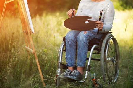 Detailed Shot Of Disabled Womans Legs. Lower Part Of Woman Body In A Wheelchair. Horizontal Picture Of Woman In A Wheelchair Holding A Palette And A Brushの写真素材