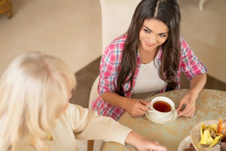 Elevated View Of The Pretty Daughter Who Attentively Listens To Her Senior Mother While Sitting At The Table And Drinking Tea.の写真素材