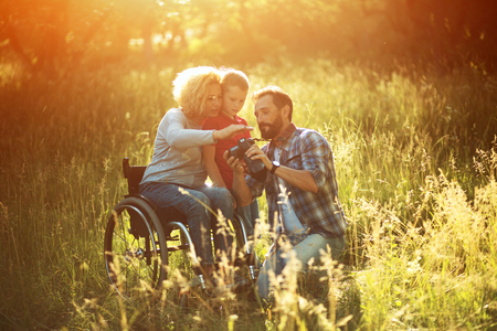 Blonde Paraplegic Woman In An Armchair Hugs Little Boy Who Stands Near Her And Looks On The Photos On Camera. Photographer Demonstrates The Result Of His Work To Disables Woman And Her Son. Outdoorsの写真素材