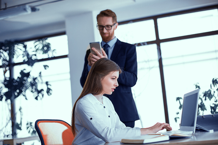 Busy girl sitting at the table and typing on a laptop. Businessman in the background is looking into the smartphone typing messageの写真素材