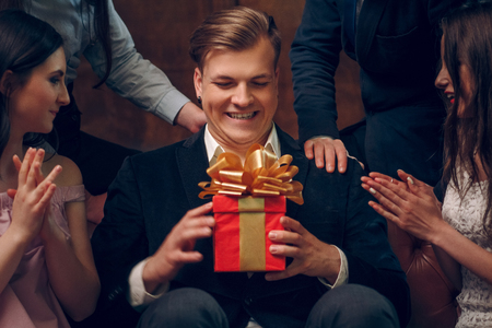 Excited man holding a birthday present from his friends at the birthday party. Smiling young man in suit sits on a couch and holds his gift for the birthday - a red box with golden ribbon. Close up shotの写真素材