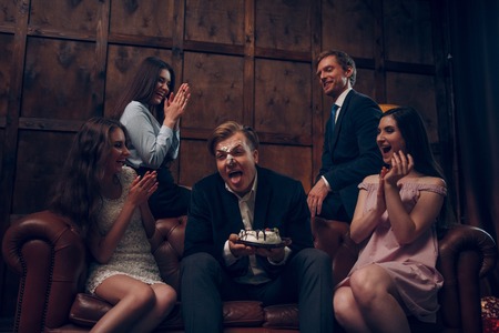 Laughing hard at young man whose face is covered with piece of birthday cake. Man whose face is in cake sticks his tongue to lick his lipsの写真素材