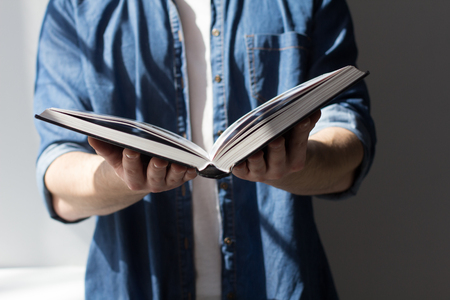 Young male holding huge book in hands. Cropped photo of guy wearing white tshirt underneath denim...