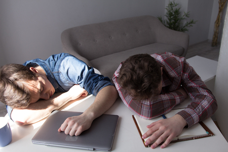 Two teenages sleeping on desk. Young teenage boys fallen asleep after studying for very long time. Living room interior in background.の写真素材